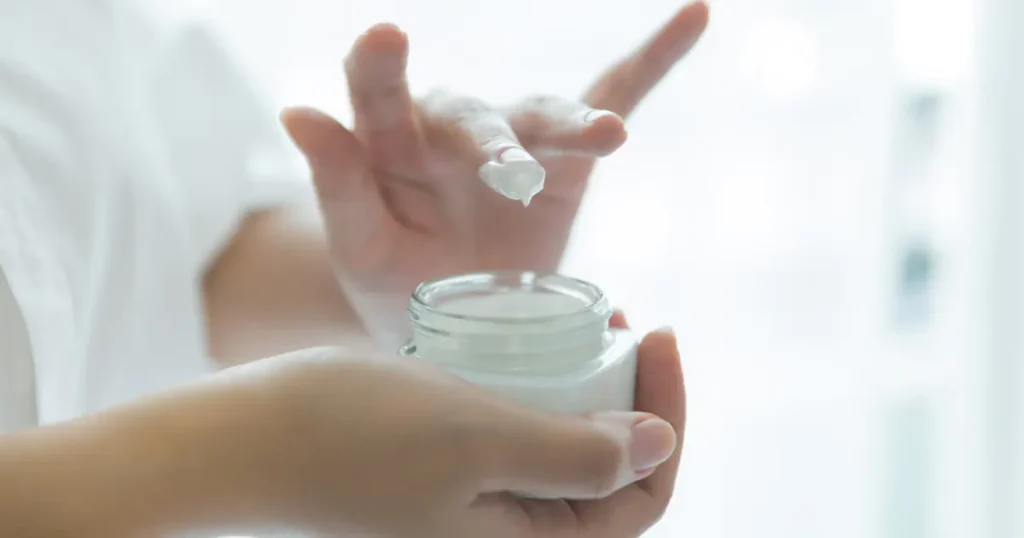 A person's hands holding a glass jar of white facial cream and taking a small amount on their fingertip against a bright, blurred background.