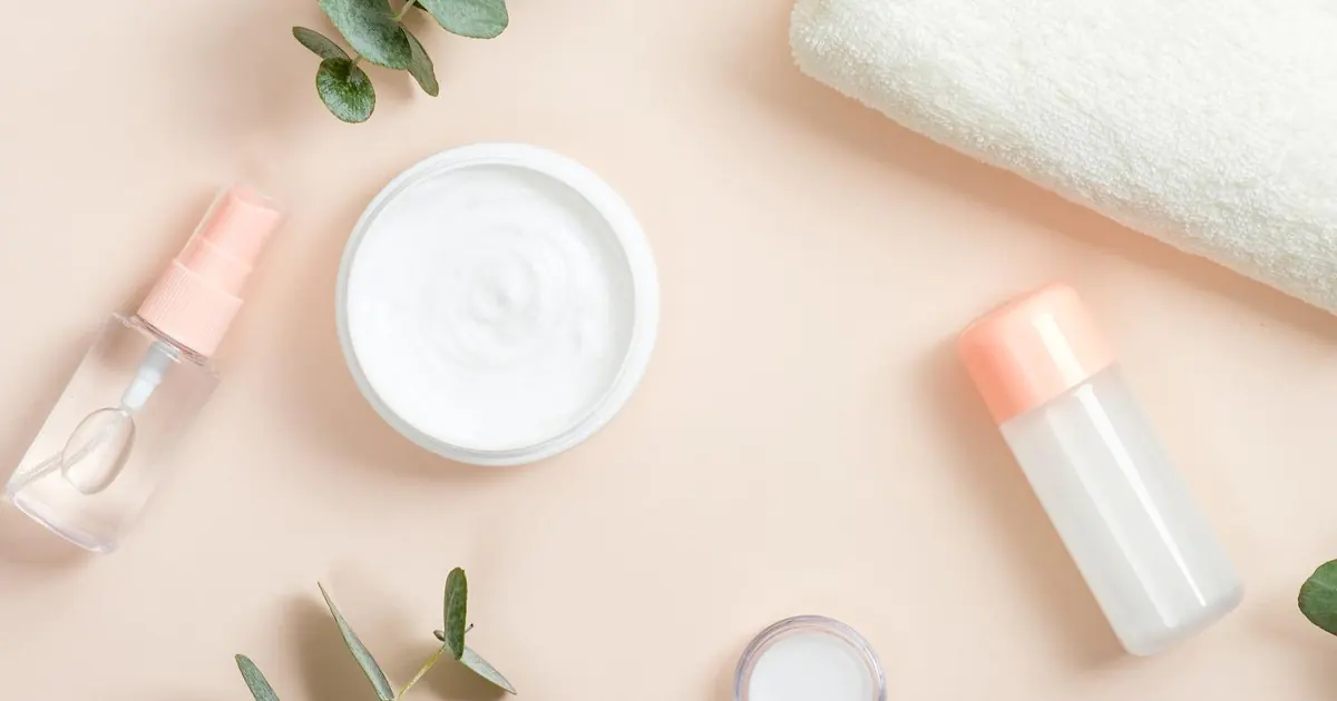 Top-down view of white skincare cream in a round jar, a pink spray bottle, and a clear serum bottle arranged on a beige surface with green eucalyptus leaves and a white towel.