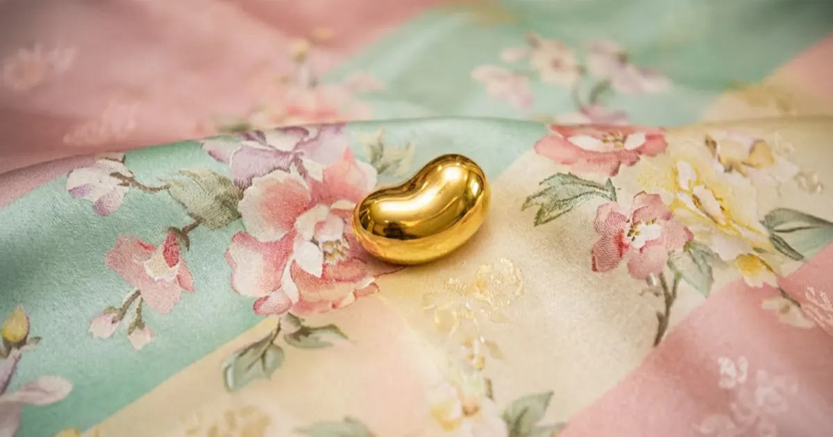 A small, smooth 24k gold bean-shaped charm resting on a colorful, floral-patterned traditional Korean silk fabric (bojagi).