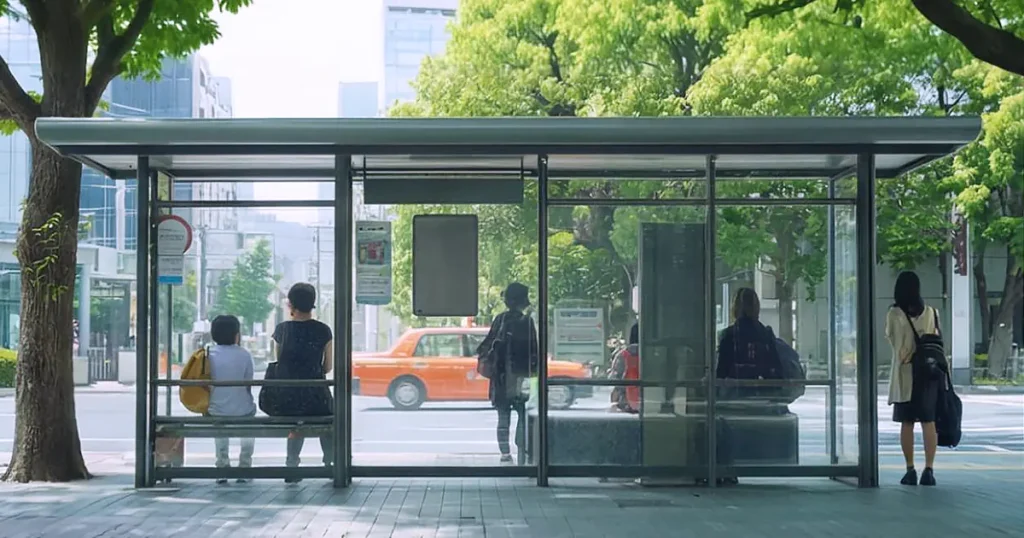 People sitting and standing at a modern glass bus stop in a city with green trees and buildings in the background.