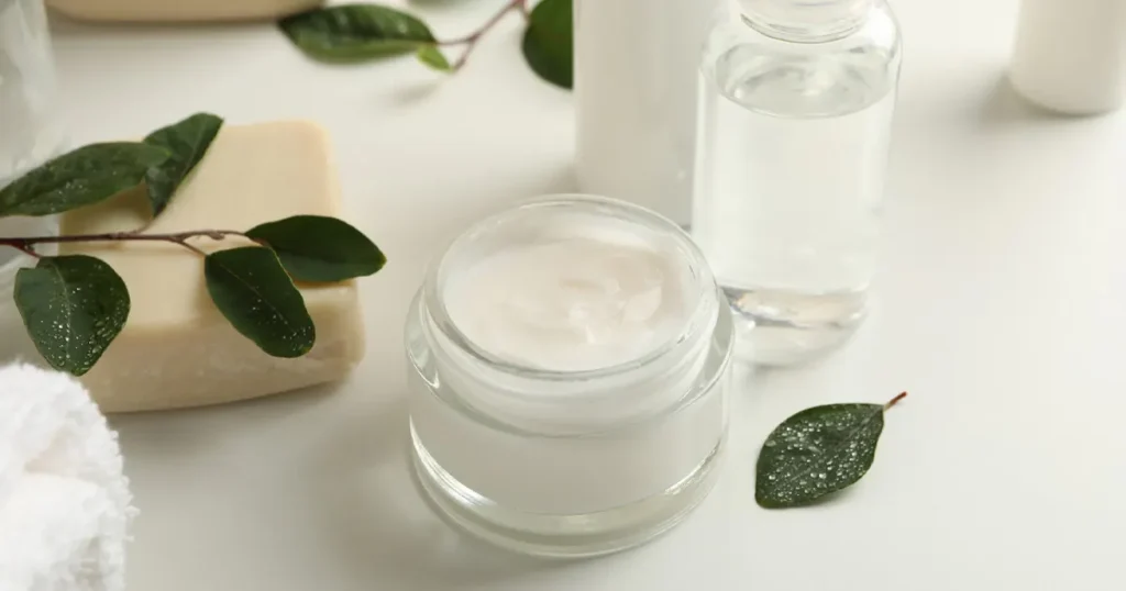 A jar of white cream, a bar of soap, a clear bottle of liquid, and green leaves with water droplets on a white surface.