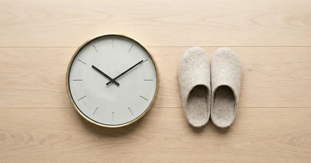 A wall clock showing time and a pair of gray felt slippers neatly placed on a light-colored wooden floor