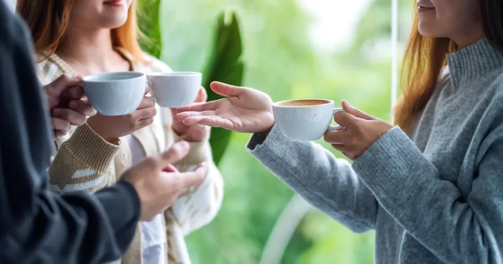 Several people standing together indoors, holding cups of coffee and gesturing as they talk.
