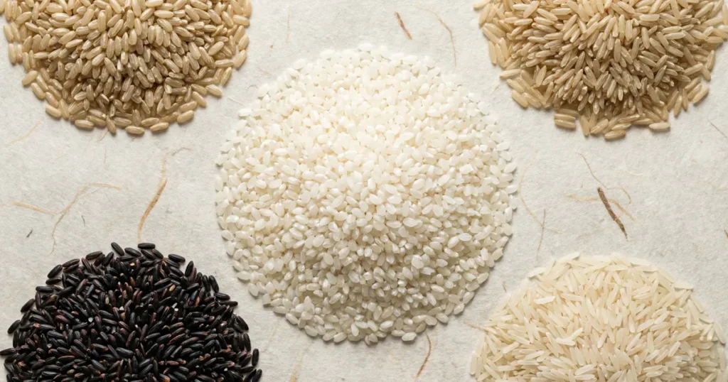 Five different heaps of rice including black rice, brown rice, and short-grain white rice arranged symmetrically on light beige traditional paper.