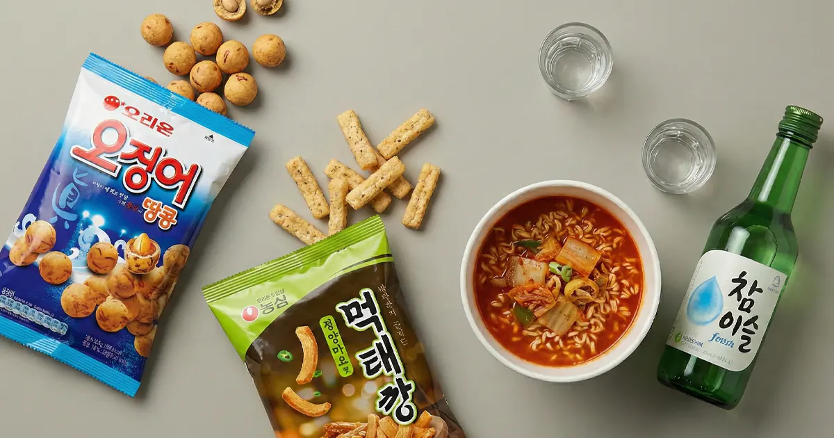 A flat lay of various Korean convenience store snacks including squid peanut crackers, pollack snacks, kimchi ramen, and a bottle of soju with shot glasses on a neutral background.