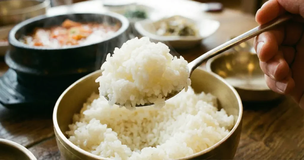 A close-up of a metal spoon lifting white rice from a brass bowl, with a steaming jjigae stew blurred in the background.