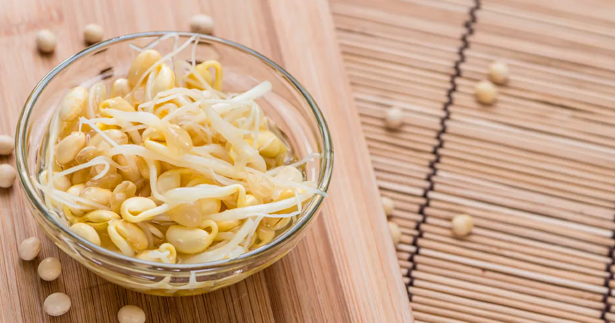 A glass bowl filled with blanched yellow soybean sprouts sits on a light wooden cutting board, surrounded by dry yellow soybeans on a bamboo mat.