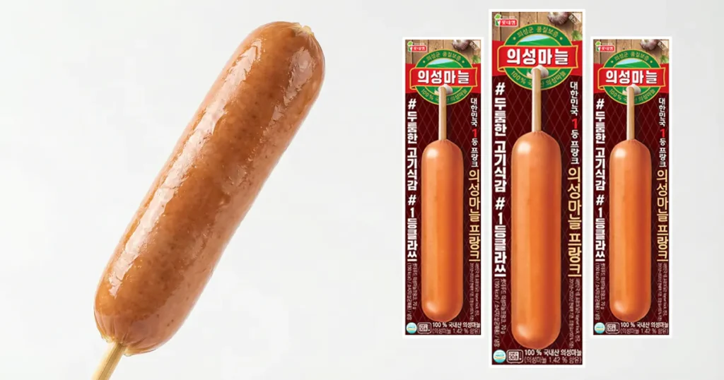 A single juicy sausage on a wooden stick next to three packages of Lotte Uiseong Garlic Hot Bar.