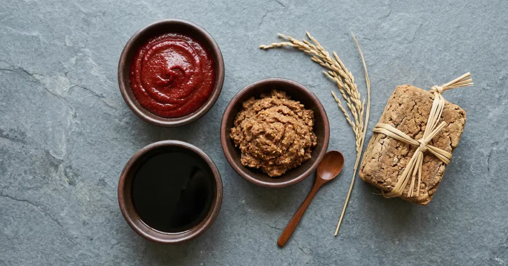 Three clay bowls containing red chili paste, dark soy sauce, and thick soybean paste are arranged next to a Meju brick on a grey stone surface.