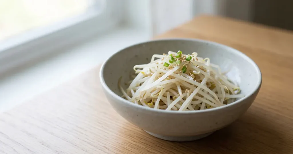 Pale yellow mung bean sprouts seasoned with sesame seeds and green onions in a light grey bowl.